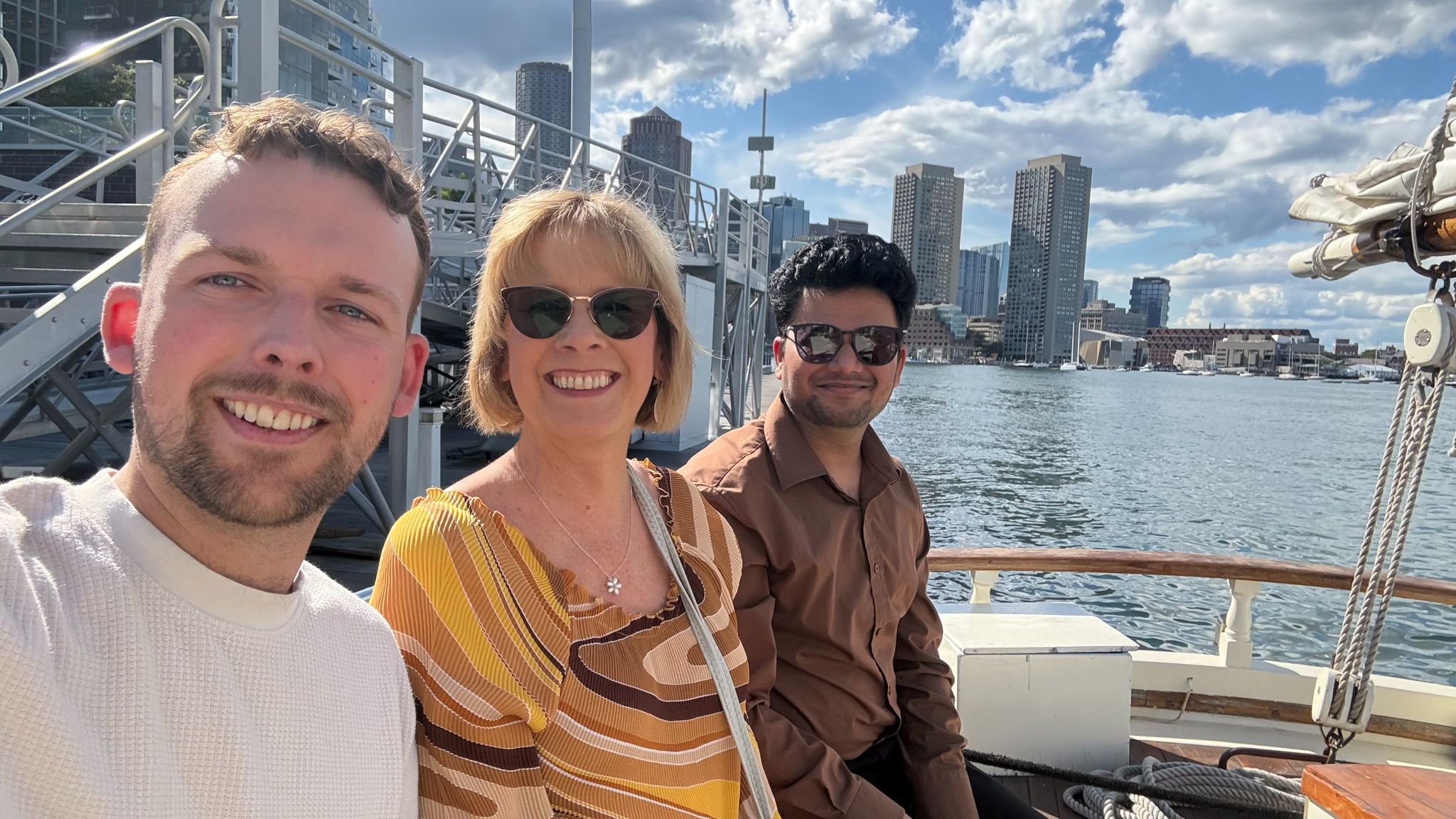Three people on a sailboat with the Boston skyline behind them